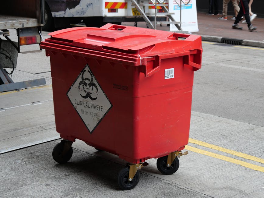 A red wheeled clinical waste bin made of durable plastic with a textured finish, positioned on a concrete pavement near a street curb. The bin has a hinged lid with a slightly raised handle for easy o