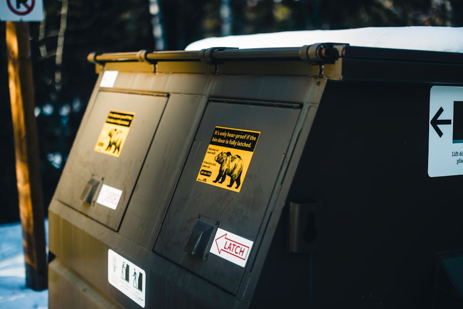 A red wheeled clinical waste bin made of durable plastic with a textured finish, positioned on a concrete pavement near a street curb. The bin has a hinged lid with a slightly raised handle for easy o