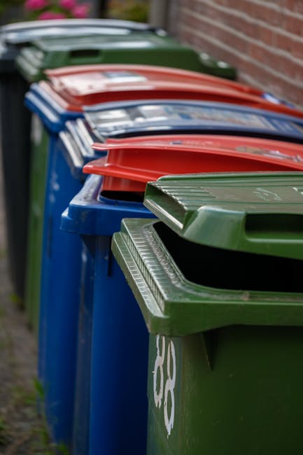 A row of large outdoor rubbish bins lined up along a pavement, with green and blue bins in the foreground and red bins further back. The green bin in the front has a hinged lid with a textured surface